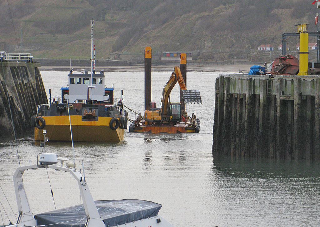 A digger fixed to a floating platform in the entrance of a harbour with an additional boat for both piloting it and taking the sediment.